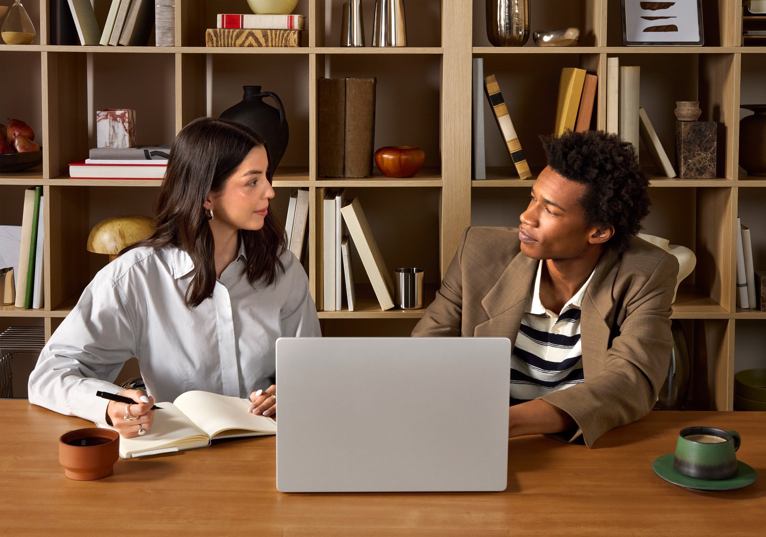 A man and woman sit at a wooden table having a conversation. The woman holds a pen and notebook, and there is a silver laptop in front of the man. The background features a wooden shelf with books and decorative items.