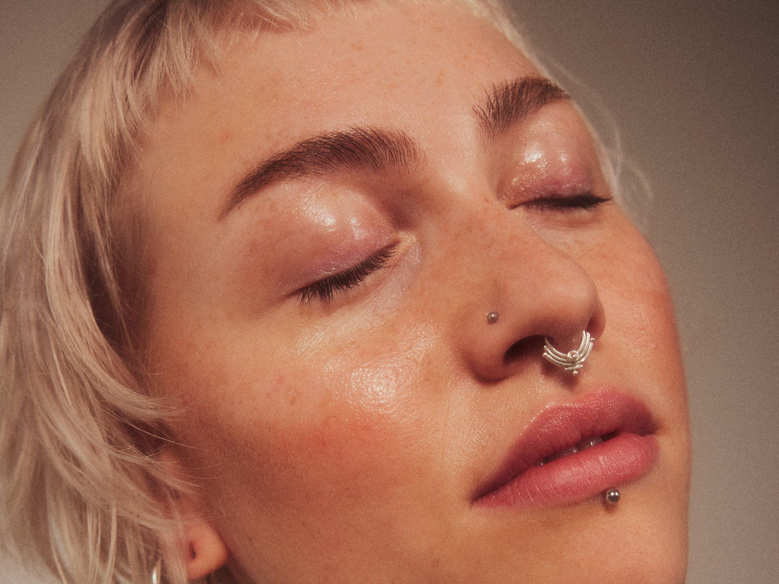 Nahaufnahme eines Models mit Piercings im Gesicht, geschlossene Augen, kurze blonde Haare, natürliche Haut, Weichzeichnung, warmes Licht.