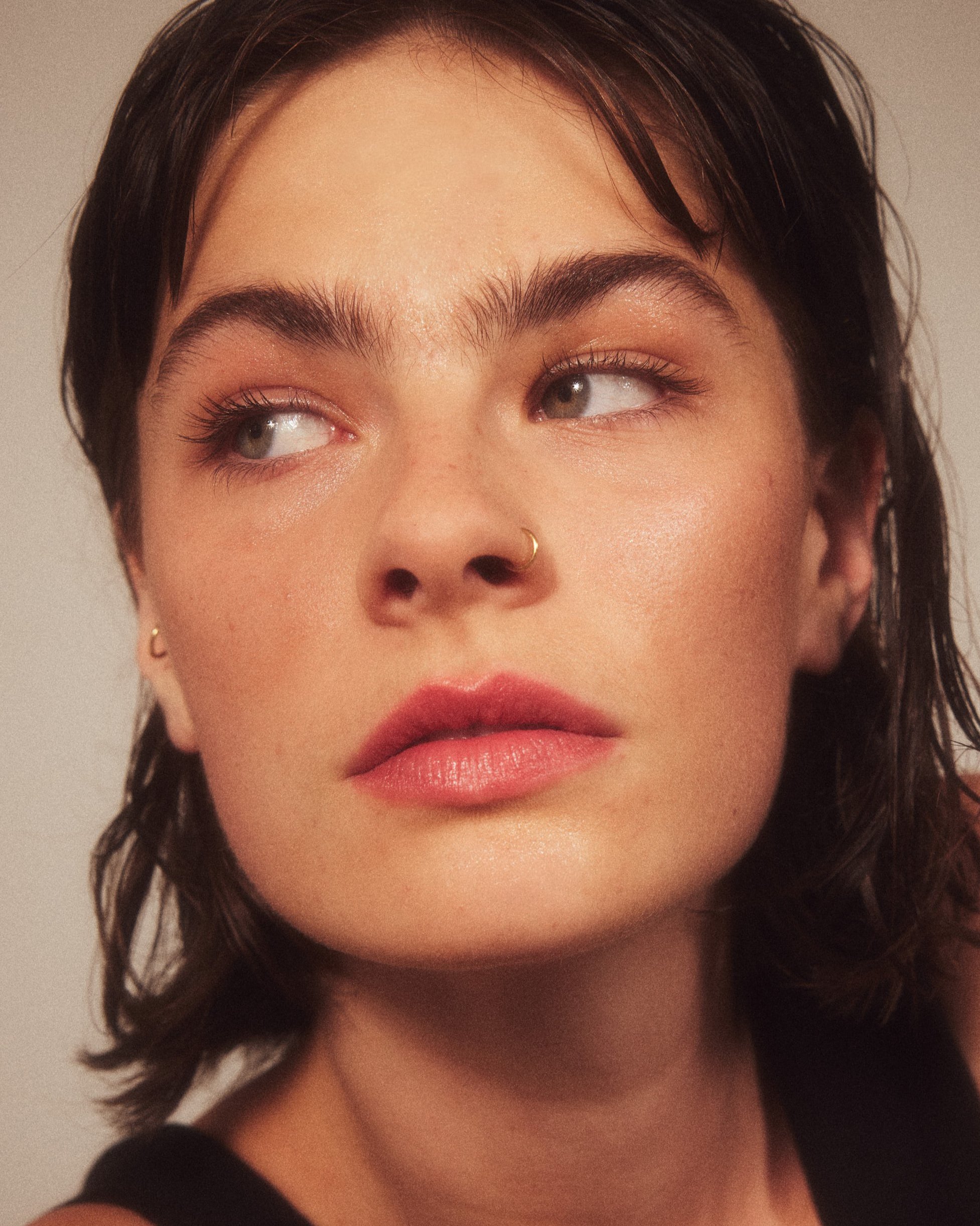 Ein junges Frau mit kurzen, dunklen Haaren, Goldohrringen und Nasenring, schaut nach links mit leicht geöffneten Lippen.