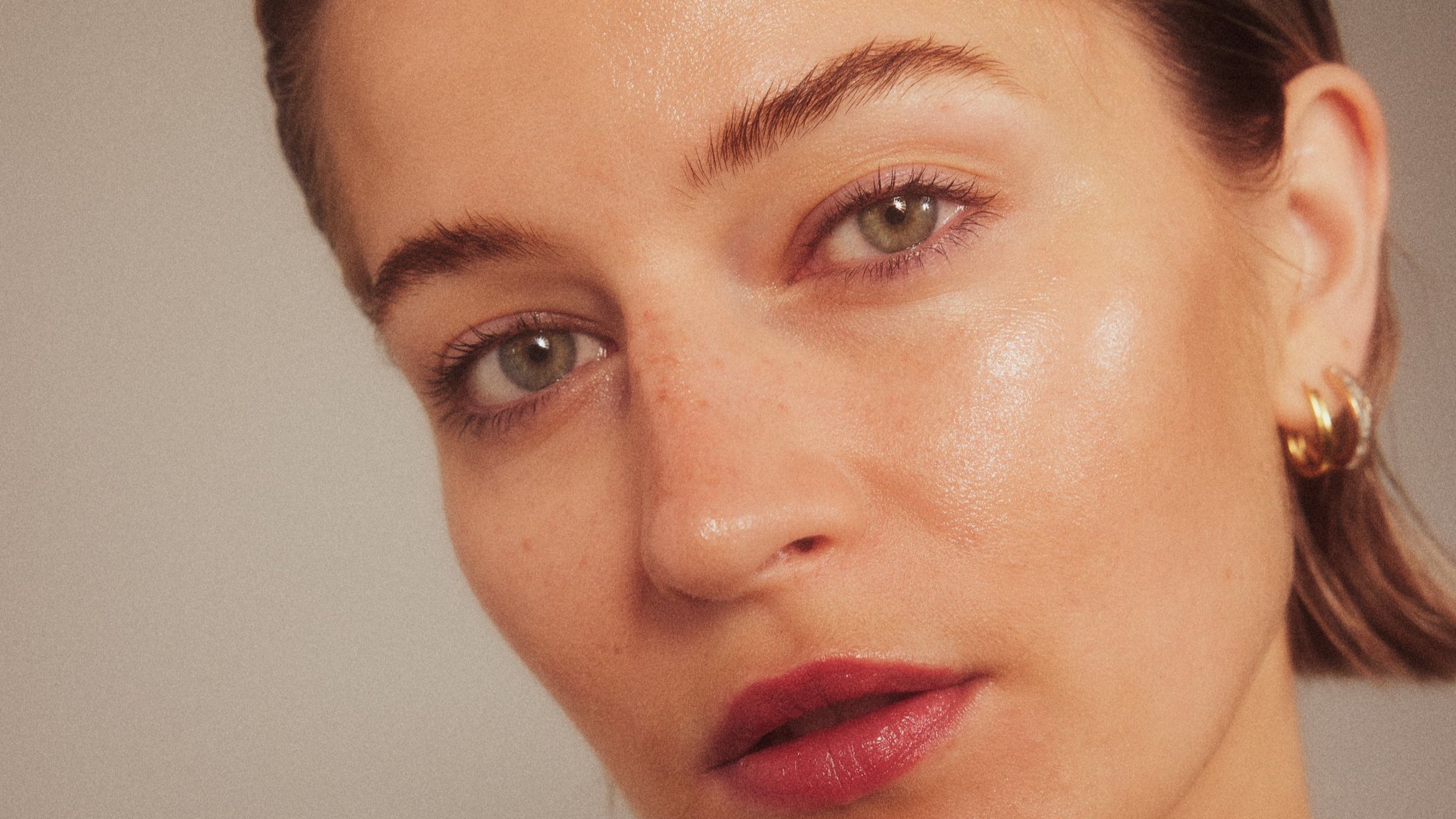 Nahaufnahme eines jungen Frauengesichts mit braunen Haaren, goldenen Ohrringen, natürlichem Make-up und leicht glühender Haut.